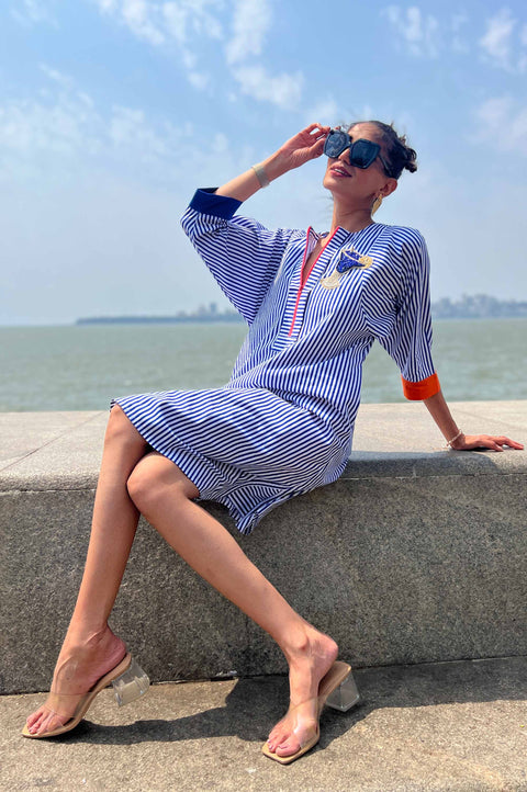 Cocktail blue and white striped dress for women with neon zipper, embroidered patch and orange pockets, seated pose by the sea