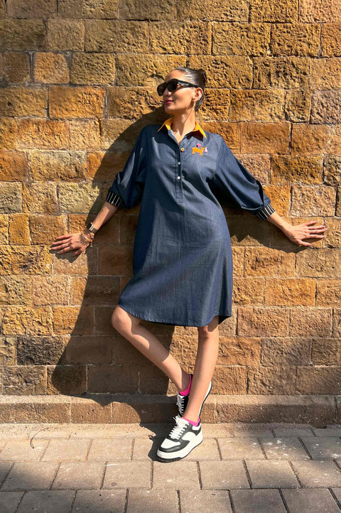 Bambi blue denim shirt dress for women with orange collar, striped cuffs and embroidered deer, leaning on a wall
